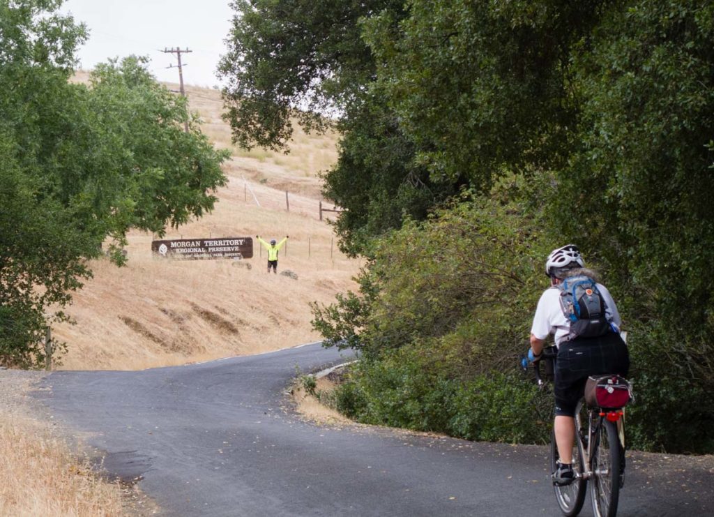 A woman on a bike rides up a rural road towards a park entrance which reads "Morgan Territory Regional Preserve." Another cyclist waits at the entrance with her hands raised triumphantly. The landscape of the park is dry grassland; on the other side of the road are dense woods.