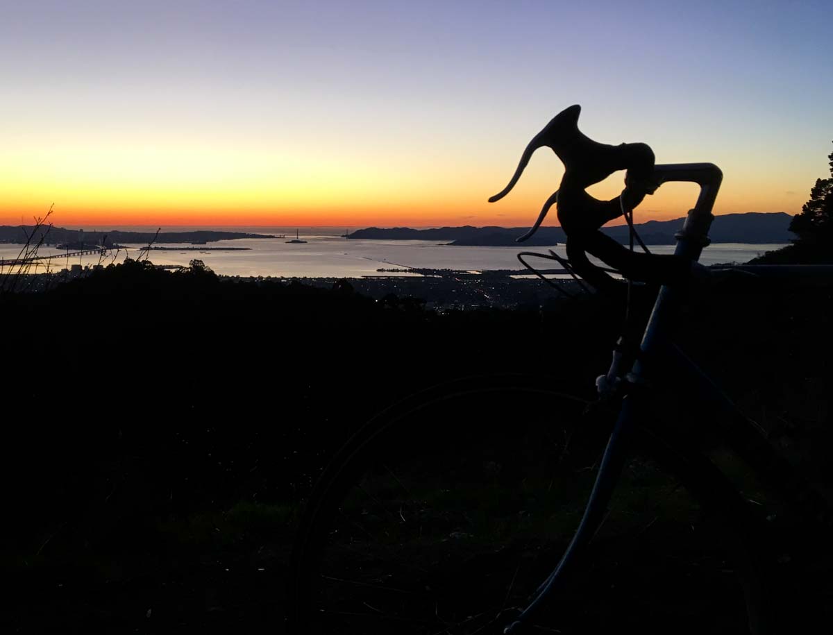 The handlebars and front fork of a road bike are silhouetted against a gradient sky at dusk. The viewpoint is at the top of a hill facing the San Francisco Bay and the Golden Gate Bridge.
