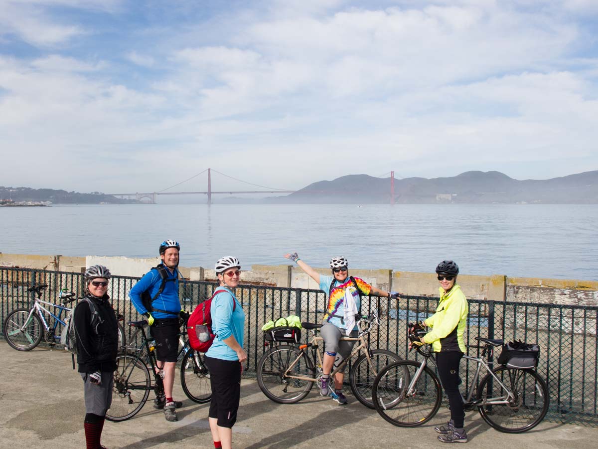 A group of cyclists stands at a seawall with the Golden Gate Bridge in the background on a misty morning. They are all smiling. One is pointing to the bridge.