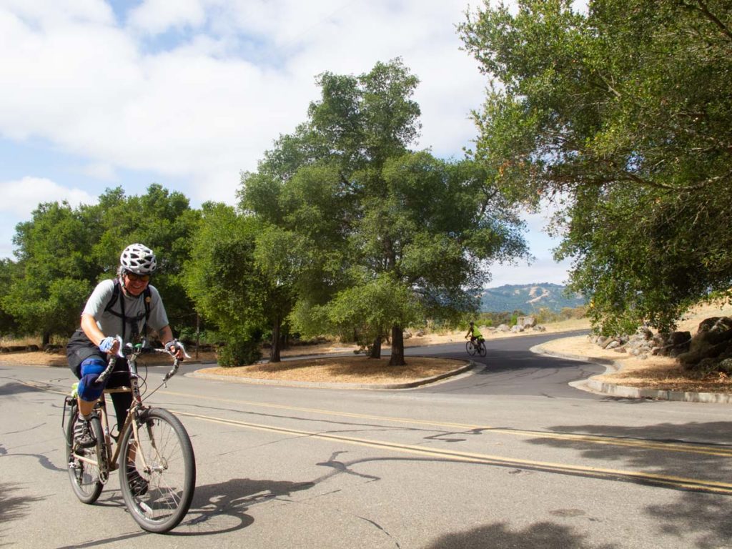 A smiling bicyclist climbs a hill on a quiet road. Oak trees line the other side of the road, and a valley and another ridge are visible in the background.